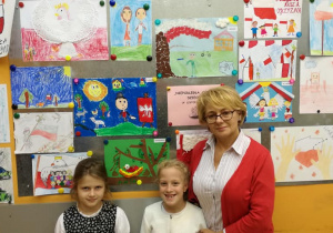 Uczennice Ania i Natalka stoją wraz z bibliotekarką Anną Załuską przy galerii prac plastyczny, które zostały nagrodzone w konkursie "Niepodległa oczami dziecka"